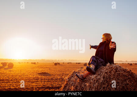 Felice di mezza età donna seduta sul pagliaio nel campo d'autunno con le braccia aperte. Rilassante e ammirando la natura al tramonto Foto Stock