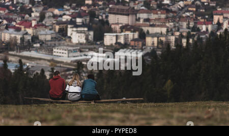 Tre amici seduti su una panca in legno che si affaccia su una piccola città Foto Stock