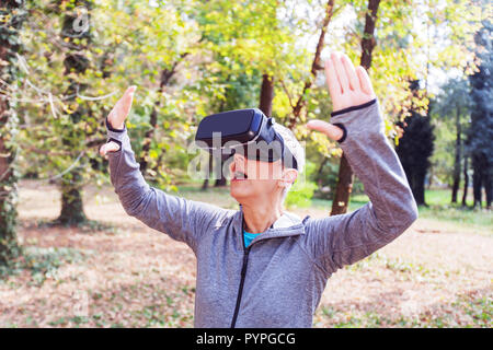 Sorpreso Senior donna divertente con la realtà virtuale auricolare in foresta, VR dispositivo utilizzando le persone anziane. Foto Stock