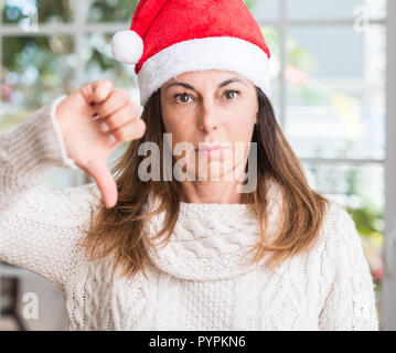 Donna di mezza età che indossa Berretto di Babbo Natale a casa con il viso arrabbiato, segno negativo che mostra antipatia con il pollice verso il basso, il concetto di rifiuto Foto Stock