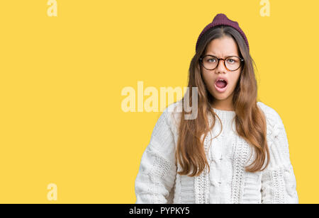 Giovane bella bruna hipster donna che indossa occhiali e cappello invernale su sfondo isolato in shock viso, cercando scettici e sarcastico, surpris Foto Stock