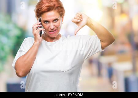 Accattivante senior redhead caucasica donna parlando sullo smartphone su sfondo isolato con volto arrabbiato, segno negativo che mostra antipatia con i pollici fare Foto Stock