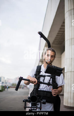 Videografo professionista tenere la fotocamera su 3 assi sospensione cardanica che montato su easy rig. Videografo utilizzando steadicam. Attrezzatura Pro aiuta a rendere i video di alta qualità senza agitazione. Foto Stock