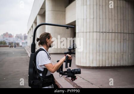 Videografo professionista tenere la fotocamera su 3 assi sospensione cardanica che montato su easy rig. Videografo utilizzando steadicam. Attrezzatura Pro aiuta a rendere i video di alta qualità senza agitazione. Foto Stock