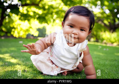 Carino bambina con un sorriso sul suo viso, arrivando a prendere il suo primo passo ridotta su un verde prato di un parco all'aperto, di etnia miste gara. Foto Stock