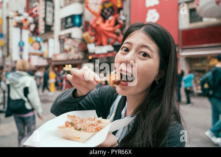 Signora giovane viaggiatore takoyaki mangiare sulla strada brulicante. ragazza cercando turistica giapponese gustosi polpo palle sulla famosa strada dotonbori. Auto-guide Foto Stock