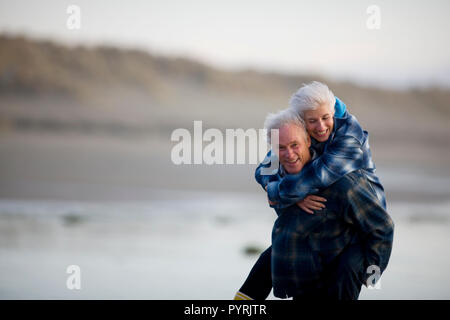 Ritratto di un sorridente uomo maturo piggybacking sua moglie lungo una spiaggia remota. Foto Stock