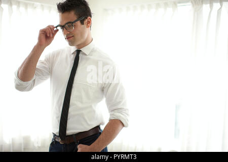 Giovane uomo che indossa una camicia e cravatta regolando i suoi occhiali mentre in una stanza con le tende tirate. Foto Stock
