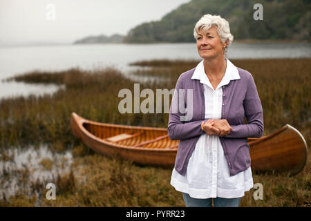 Donna matura sembra lontano come lei sta con le mani giunte in posa per un ritratto di fronte una canoa di legno su un lago. Foto Stock