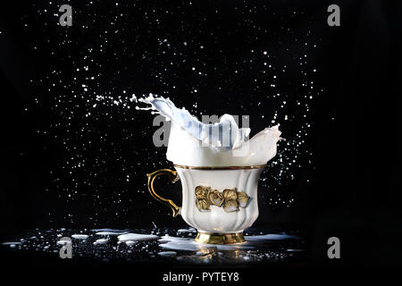 Abbondante latte schizza volare fuori della proteina mug con un manico oro e un fiore su uno sfondo nero. Foto Stock