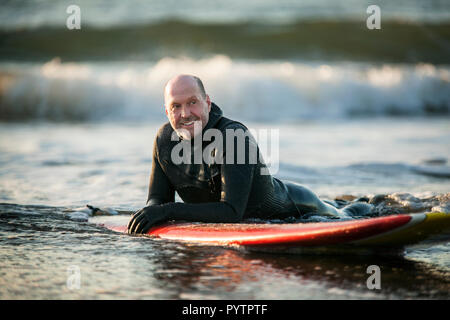 Sorridente metà uomo adulto in una muta galleggiante sulla sua tavola da surf in mare. Foto Stock