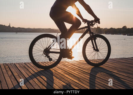 Uomo Bicicletta Equitazione al tramonto, escursioni in bicicletta in estate, silhouette del ciclista vicino al lago Foto Stock