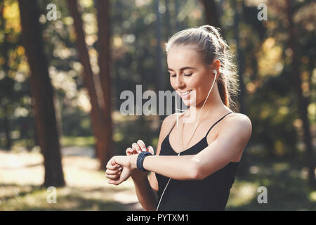 Sportish dai lunghi capelli biondo giovane donna che indossa la maglia nera in piedi nel soleggiato parco, ascoltando musica e guardando sullo sport watch, pensa circa la perdita di calorie, uno stile di vita sano e concetto di persone Foto Stock