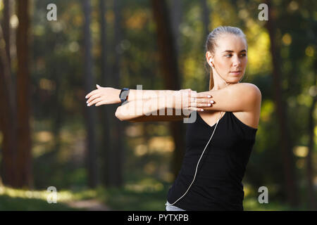 Ritratto di giovane donna atletica indossando vestiti sportish stretching e ascolto di musica nel soleggiato parco, uno stile di vita sano e concetto di persone Foto Stock