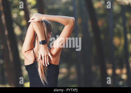 Ritratto di giovane atletico dai capelli lunghi donna che indossa abiti sportish stretching le sue braccia e ascolto di musica nel soleggiato parco, uno stile di vita sano e concetto di persone Foto Stock