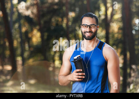 Ritratto di giovane atletico uomo multiculturale indossando occhiali sportish avente pausa e acqua potabile nel soleggiato parco, uno stile di vita sano e concetto di persone Foto Stock
