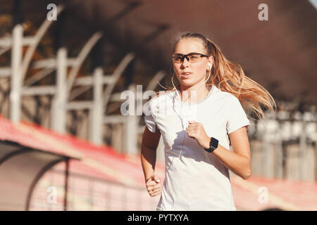 Bionda Fitness capelli lunghi donna abbigliamento sportivo in ascolto di musica e l'esecuzione in corrispondenza dello stadio di calcio all'aperto, uno stile di vita sano e concetto di persone Foto Stock