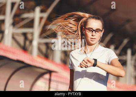 Bionda Fitness capelli lunghi donna abbigliamento sportivo in ascolto di musica e la ricerca sugli indicatori sul suo orologio sportivo mentre è in esecuzione con lo stadio di calcio all'aperto, uno stile di vita sano e concetto di persone Foto Stock