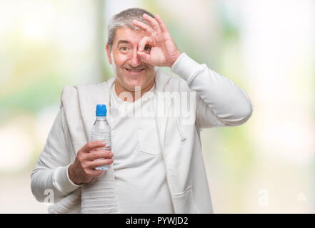 Bello il senior uomo azienda formazione asciugamano e una bottiglia d'acqua su sfondo isolato con buon viso sorridente facendo segno OK con la mano sull'occhio cercando th Foto Stock