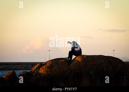 Un giovane prende un selfie mentre seduto su alcune rocce al tramonto. Spia della tenuta in caldo. Foto Stock