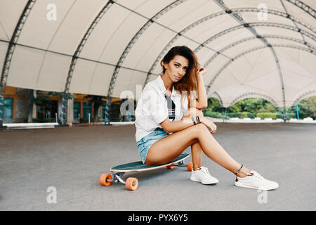 Elegante ragazza urbano in posa con Longboard. Hipster ragazza con il pattino in città. Sport estremo e concetto di emozioni Foto Stock