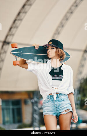 Elegante ragazza mostra atteggiamento close up ritratto, tenendo longboard sulle spalle. Sport estremo, moda giovane e concetto di emozioni Foto Stock