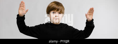Ritratto di adorabili grave little boy in piedi in studio con le mani in alto su sfondo grigio Foto Stock