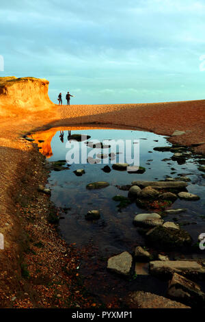 Un paio di camminare sulla spiaggia a Seatown nel Dorset vicino al tramonto con riflessi in un flusso Foto Stock