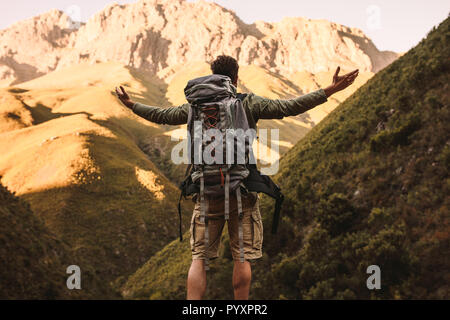 Vista posteriore del giovane uomo con zaino in piedi con le braccia aperte e guardando il paesaggio di montagna vista. Escursionista maschio sorge su una roccia e l Foto Stock