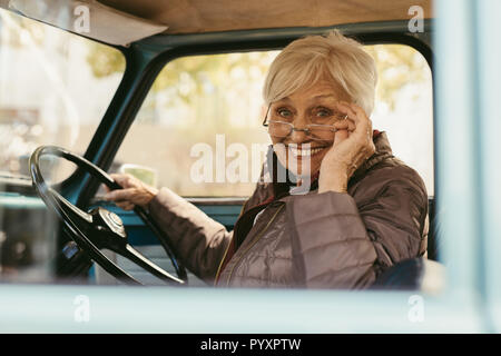 Senior donna seduta nel sedile di guida di auto tenendo gli occhiali in basso per guardare la fotocamera. Il sambuco donna alla guida di una vettura vecchia sul giorno d'inverno. Foto Stock