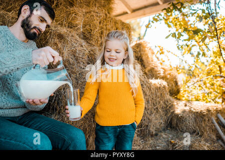 Femmina adulta agricoltore versando il latte alla piccola figlia vicino a pile di fieno al ranch Foto Stock