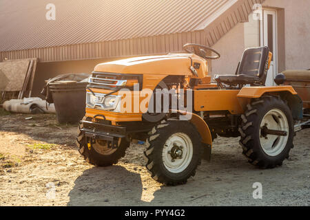 Mini trattore rosso con rimorchio in piedi vicino hangar edificio in azienda durante il tramonto o l'alba. Piccole macchine agricole. Paese rurale scena Foto Stock