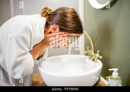 Donna in accappatoio lavando il suo volto da maschera per il viso in bagno Foto Stock