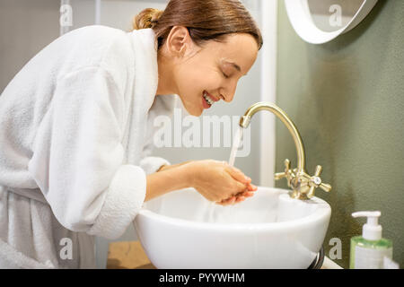 Donna in accappatoio lavando il suo volto da maschera per il viso in bagno Foto Stock