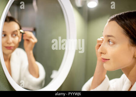 Donna in accappatoio pittura sopracciglia vicino al specchio rotondo verde sulla parete della stanza da bagno Foto Stock