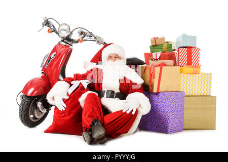 Stanco di santa claus seduto sul pavimento con rosso vintage scooter, sacco e confezioni regalo isolato su bianco Foto Stock