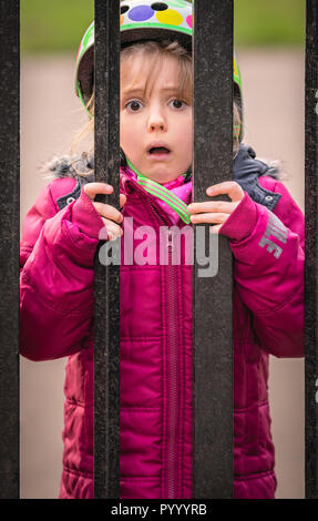 Poco carino Caucasian ragazza vestita in camicia rosa in piedi dietro i cancelli di metallo che conduce al parco giochi Foto Stock