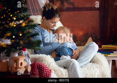La madre di allattamento al seno il suo bambino figlio seduto in poltrona accogliente vicino ad albero di Natale, inverno Foto Stock