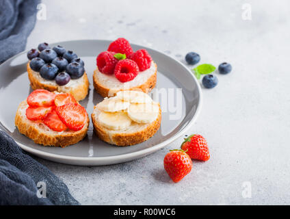 Fresco e salutare mini sandwich con crema di formaggio, frutta e bacche di lastra grigia con un panno. Fragole, mirtilli, banane e lamponi su SAN Foto Stock