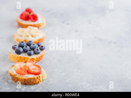 Fresco e salutare mini sandwich con crema di formaggio, frutta e bacche. Fragole, mirtilli, banane e lamponi su pietra tavolo da cucina backgro Foto Stock