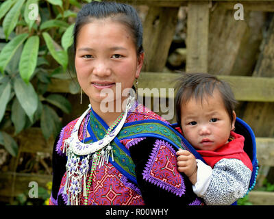 Giovane vietnamita Flower H'mongs hill-tribe donna indossa il suo clan tradizionale di un vestito ricamato e porta il suo bambino il figlio in una fionda bambino sulla schiena. Foto Stock