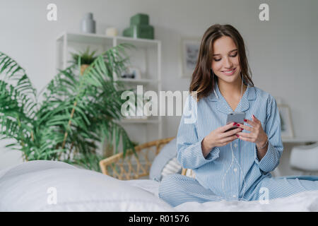 Donna felice con look accattivante, detiene il moderno telefono cellulare, Download brano alla playlist, ascolta la musica con gli auricolari, siede gambe incrociate sul confortevole b Foto Stock