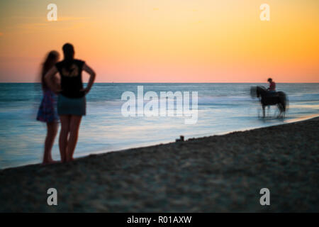 Due donne contemplando un cavaliere sulla spiaggia al tramonto, Ayamonte, Spagna. Foto Stock