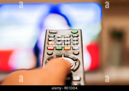 Una mano premendo un tasto del telecomando per cambiare i canali di fronte a una sfocata sullo schermo della televisione Foto Stock