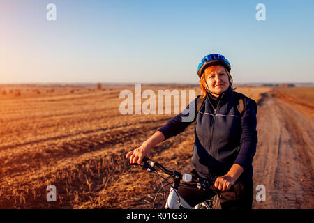 Donna di mezza età ciclista di equitazione in campo d'autunno al tramonto. Senior sportive godendo di hobby. Uno stile di vita sano Foto Stock