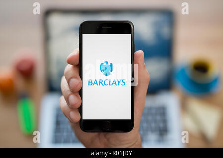 Un uomo guarda al suo iPhone che visualizza il logo di Barclays, mentre se ne sta seduto alla sua scrivania per computer (solo uso editoriale). Foto Stock