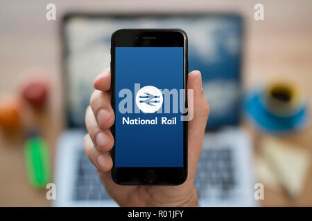 Un uomo guarda al suo iPhone che visualizza il National Rail Enquiries logo, mentre se ne sta seduto alla sua scrivania per computer (solo uso editoriale). Foto Stock