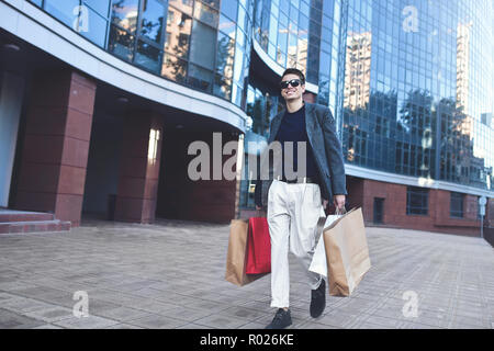 Raffreddare positivo Ragazzo con occhiali da sole e sacchi di carta camminare vicino a vetrina con la riflessione. Foto Stock