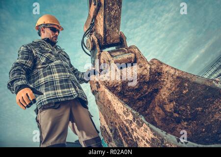 Sito in costruzione e i macchinari pesanti. Opere di terra Scavo. Lavoratore caucasica nel Casco giallo lato alla benna di un escavatore. Foto Stock
