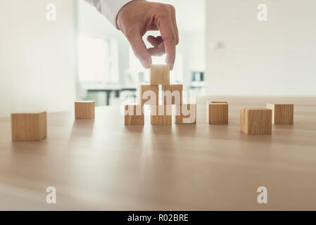 Imprenditore ponendo i cubi di legno in una forma a piramide sulla scrivania in ufficio nel suo posto di lavoro. Foto Stock